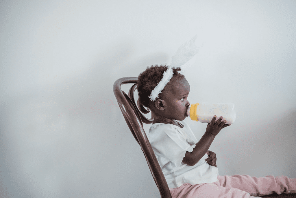 Menina pequena sentada em uma cadeira e bebendo leite da mamadeira.