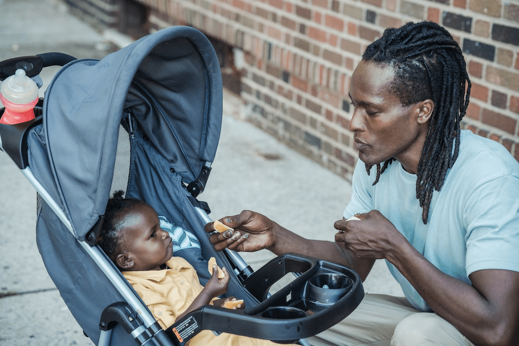 Pai alimentando filho em carrinho de bebê com 3 rodas.
