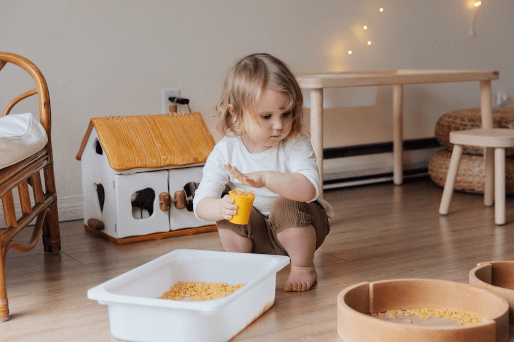 Criança segurando copo plástico amarelo cheio de macarrão, rodeada de brinquedos educativos.