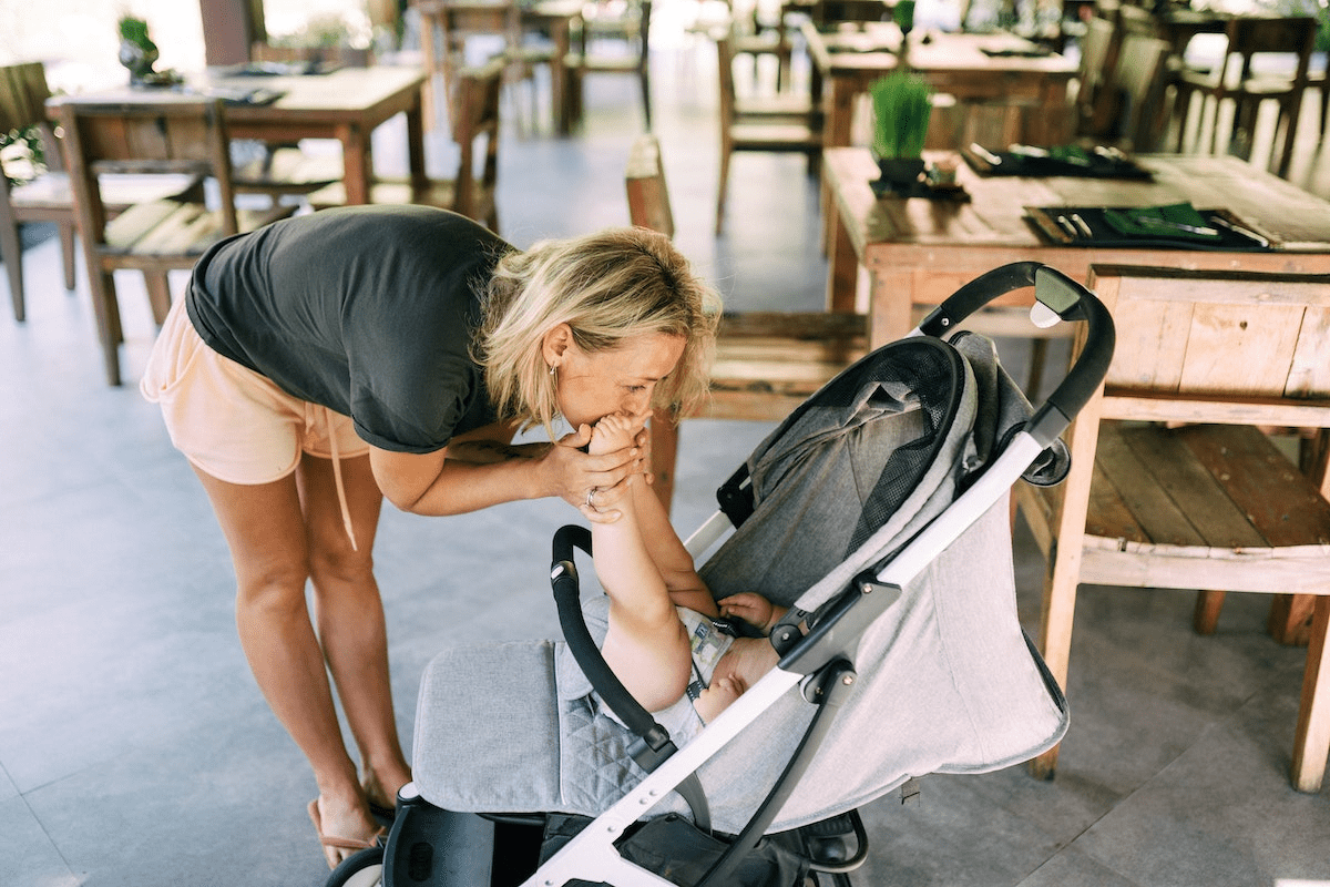 Mulher de camiseta preta e shorts inclinando-se para beijar os pezinhos de um bebê em um carrinho de passeio.