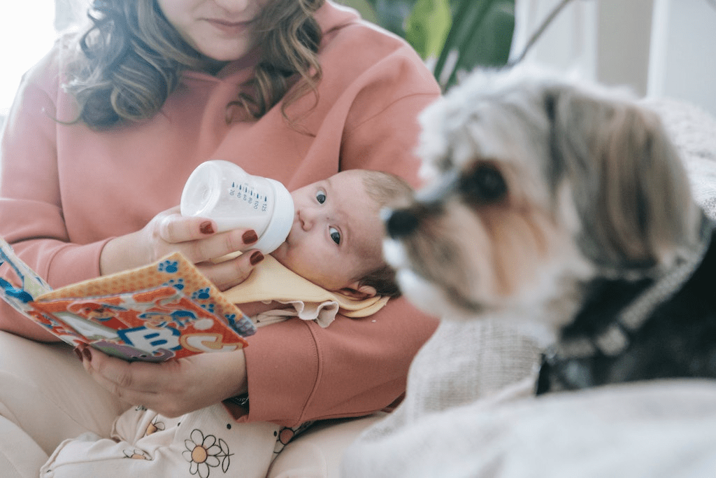 Mãe alimentando bebê recém-nascido perto de um cachorro com uma mamadeira.