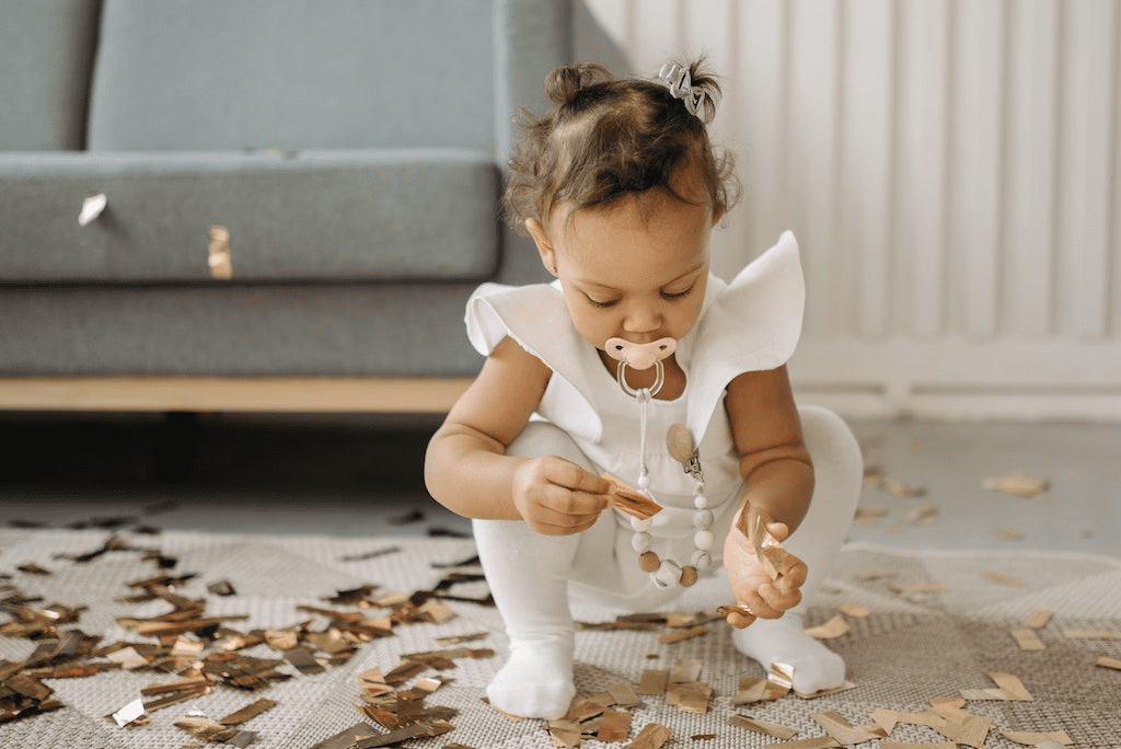 Uma criança de vestido branco e chupeta na boca pegando confete.