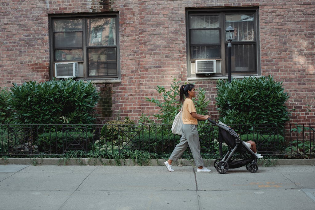 Uma mãe empurrando um carrinho de bebê pela calçada de uma rua residencial, demonstrando a rotina urbana e a mobilidade que o carrinho proporciona.